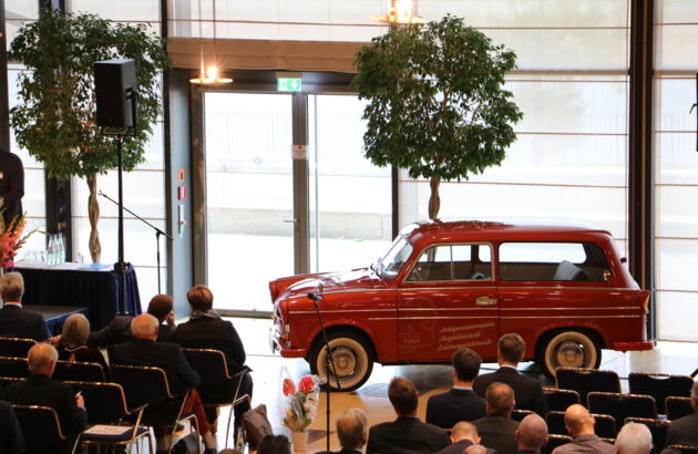 Foto einer Kon­ferenz. Zu sehen sind einige Menschen und auf der Bühen steht ein altes, rotes Auto.