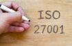 A person writes ISO_27001 on a wooden background.